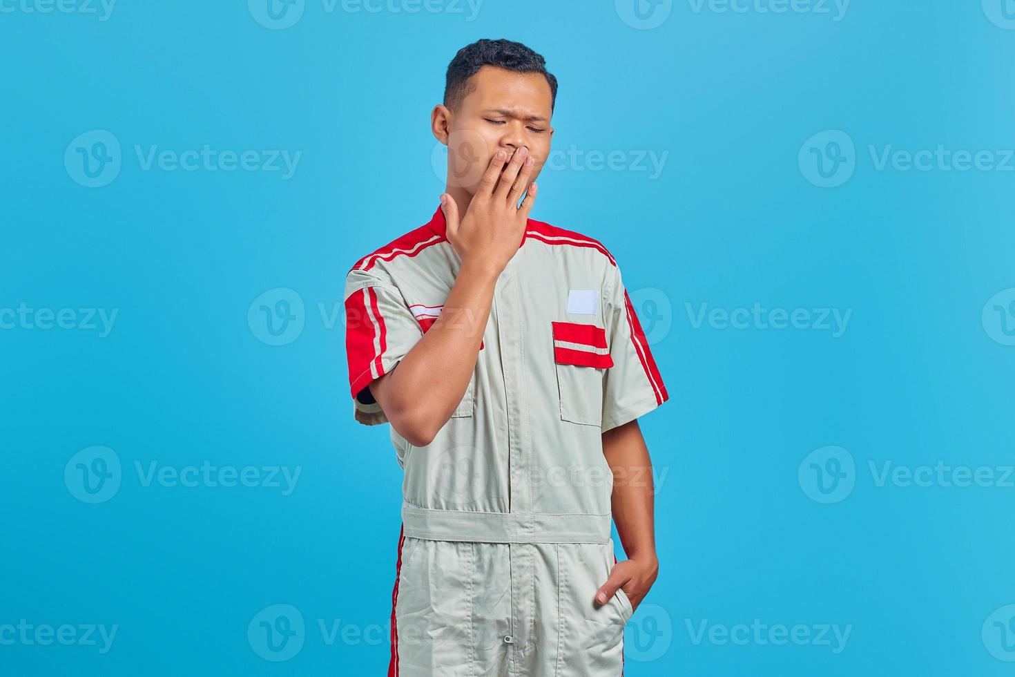 Retrato de joven mecánico sintiéndose somnoliento y tapándose la boca con la mano sobre fondo azul. foto