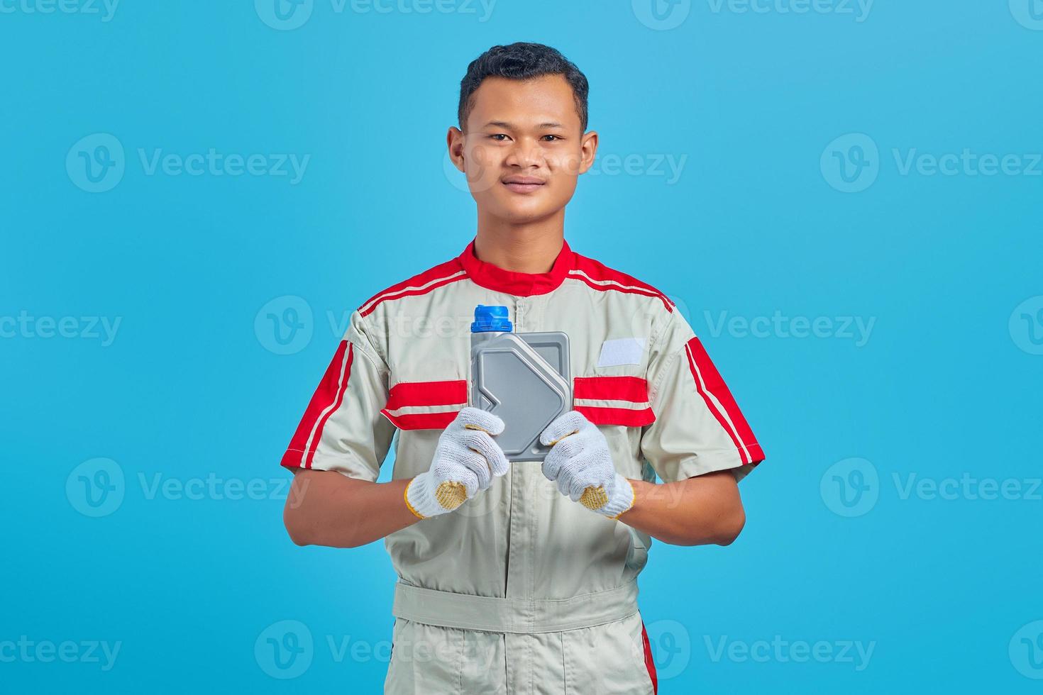 Retrato de sonriente joven mecánico asiático mostrando una botella de plástico de aceite de motor en la mano aislado sobre fondo azul. foto