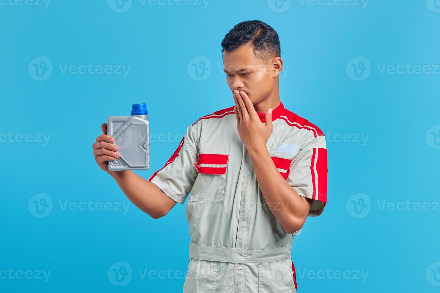 Retrato de joven mecánico asiático conmocionado sosteniendo una botella de plástico de aceite de motor y cubriendo la boca sobre fondo azul manos foto