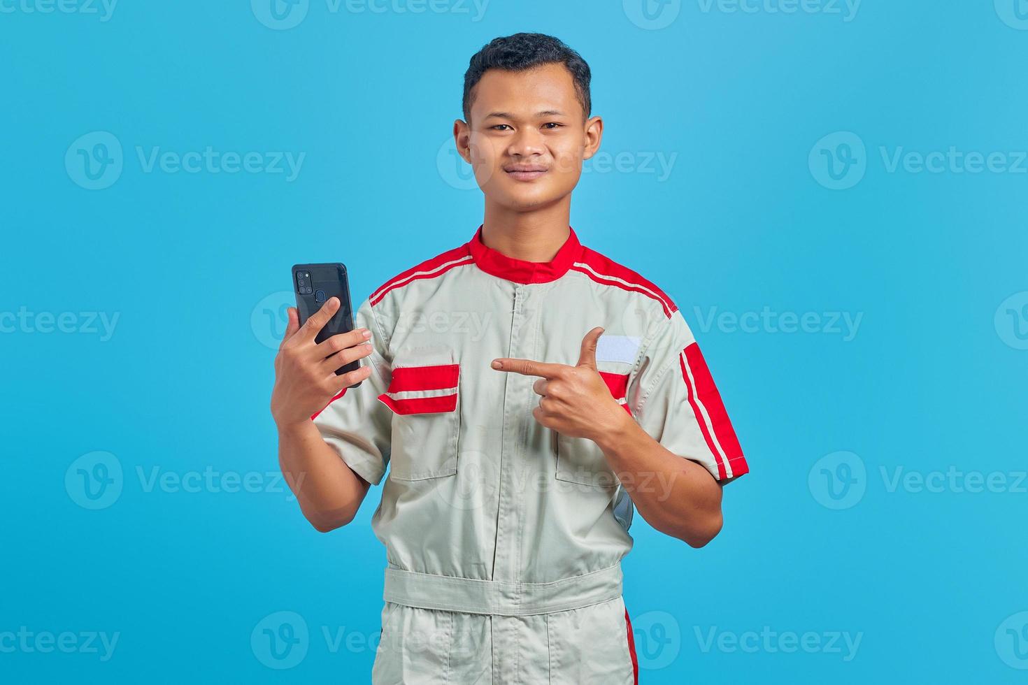 Portrait of smiling young mechanic pointing finger at mobile phone isolated on blue background photo