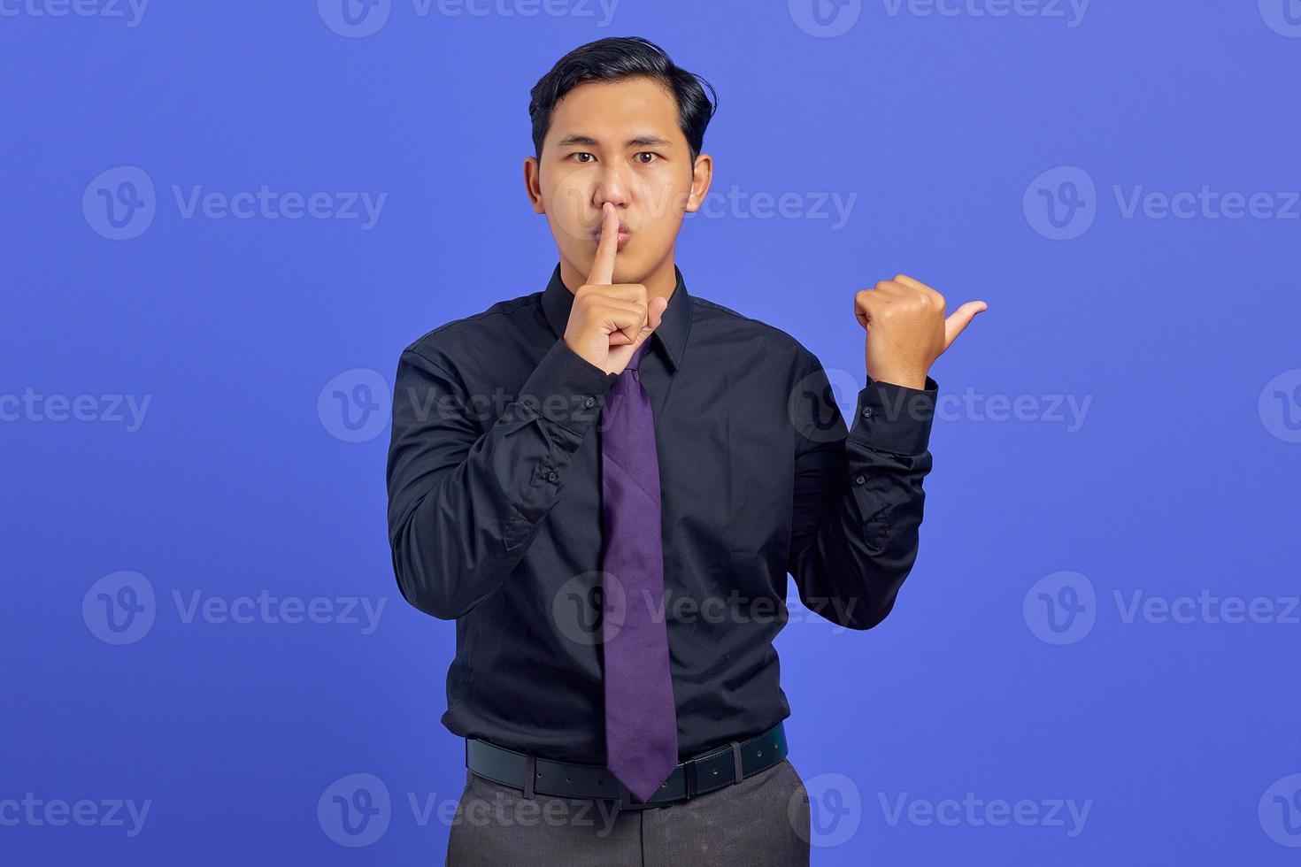 Handsome asian man signal to silence and pointing to the side over purple background photo