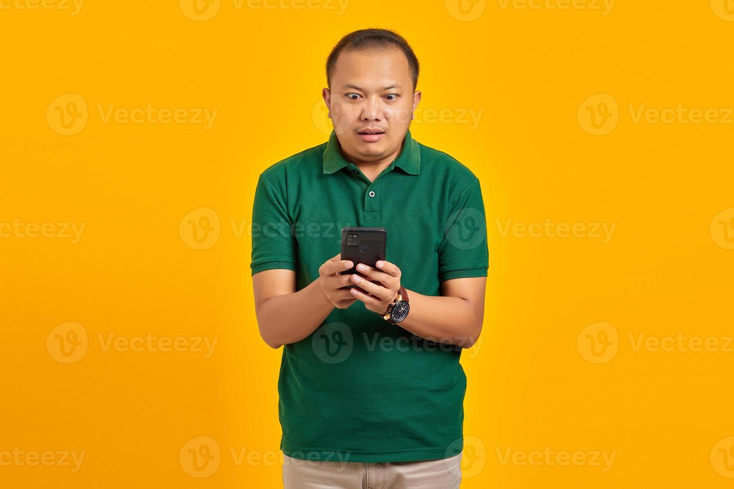 Shocked handsome young Asian man looks at smartphone screen isolated on yellow background photo