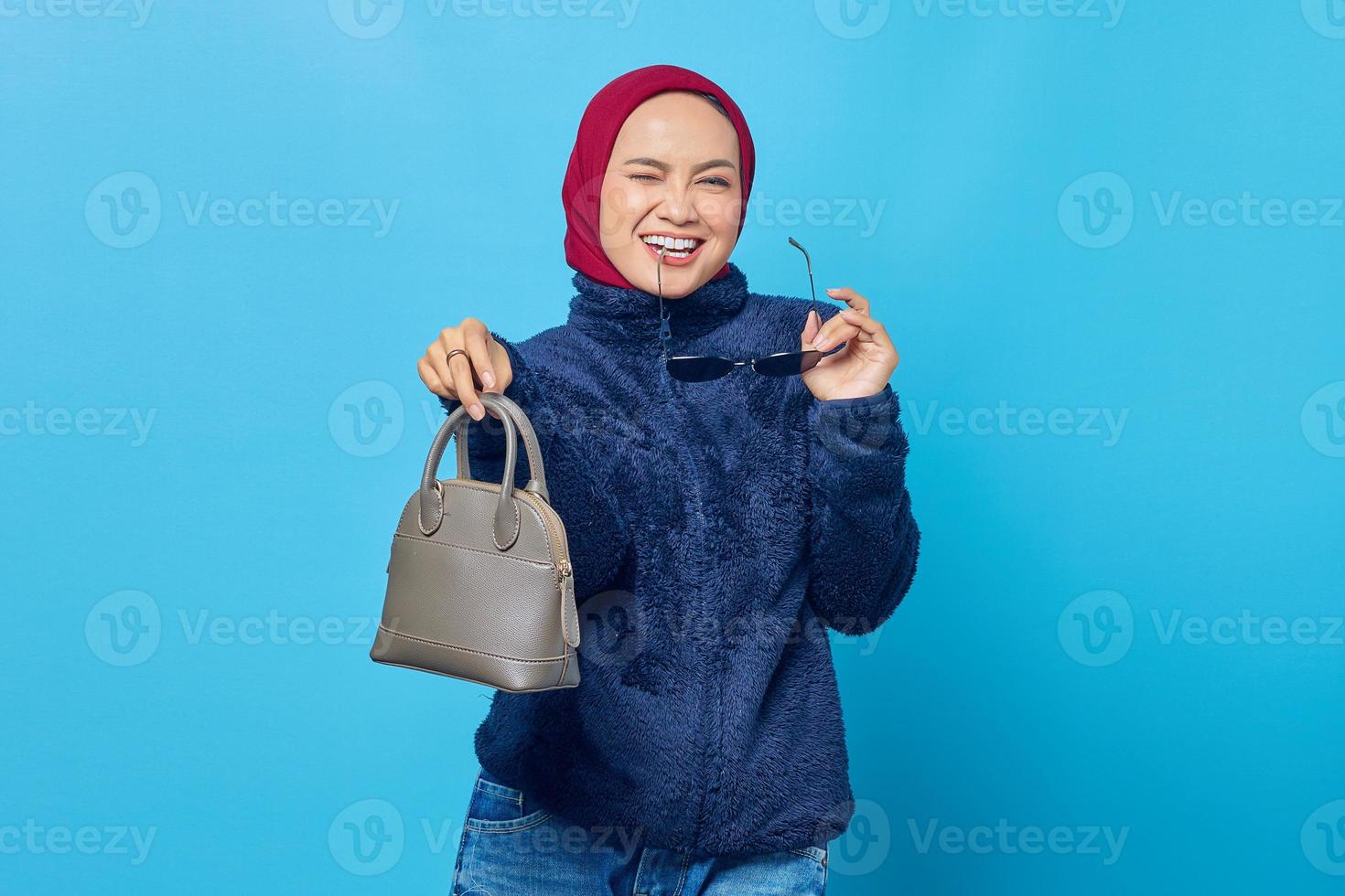 Sonriente joven mujer asiática sosteniendo la bolsa y mordiendo las gafas sobre fondo azul. foto