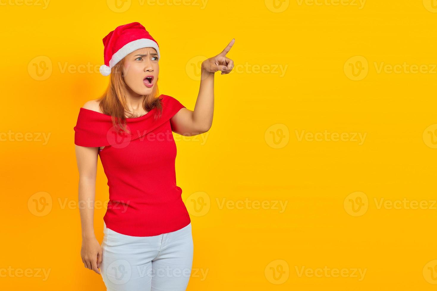 Surprised young Asian woman wearing Christmas hat and pointing sideways over yellow background photo