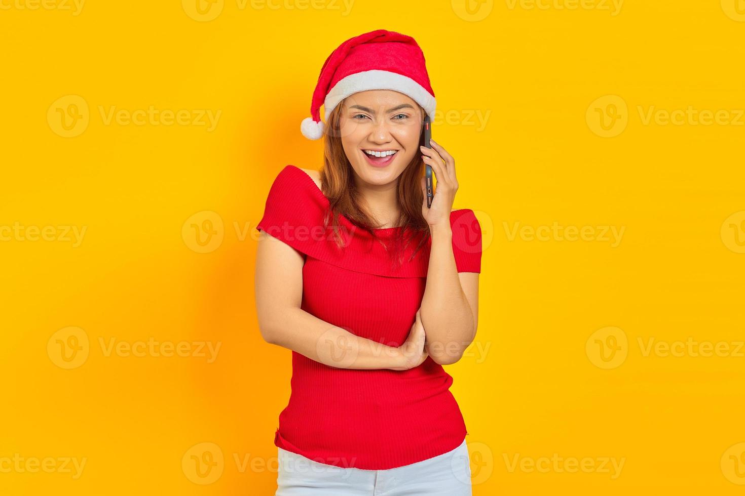 Smiling young Asian woman wearing Christmas hat talking on smartphone and looking at camera on yellow background photo