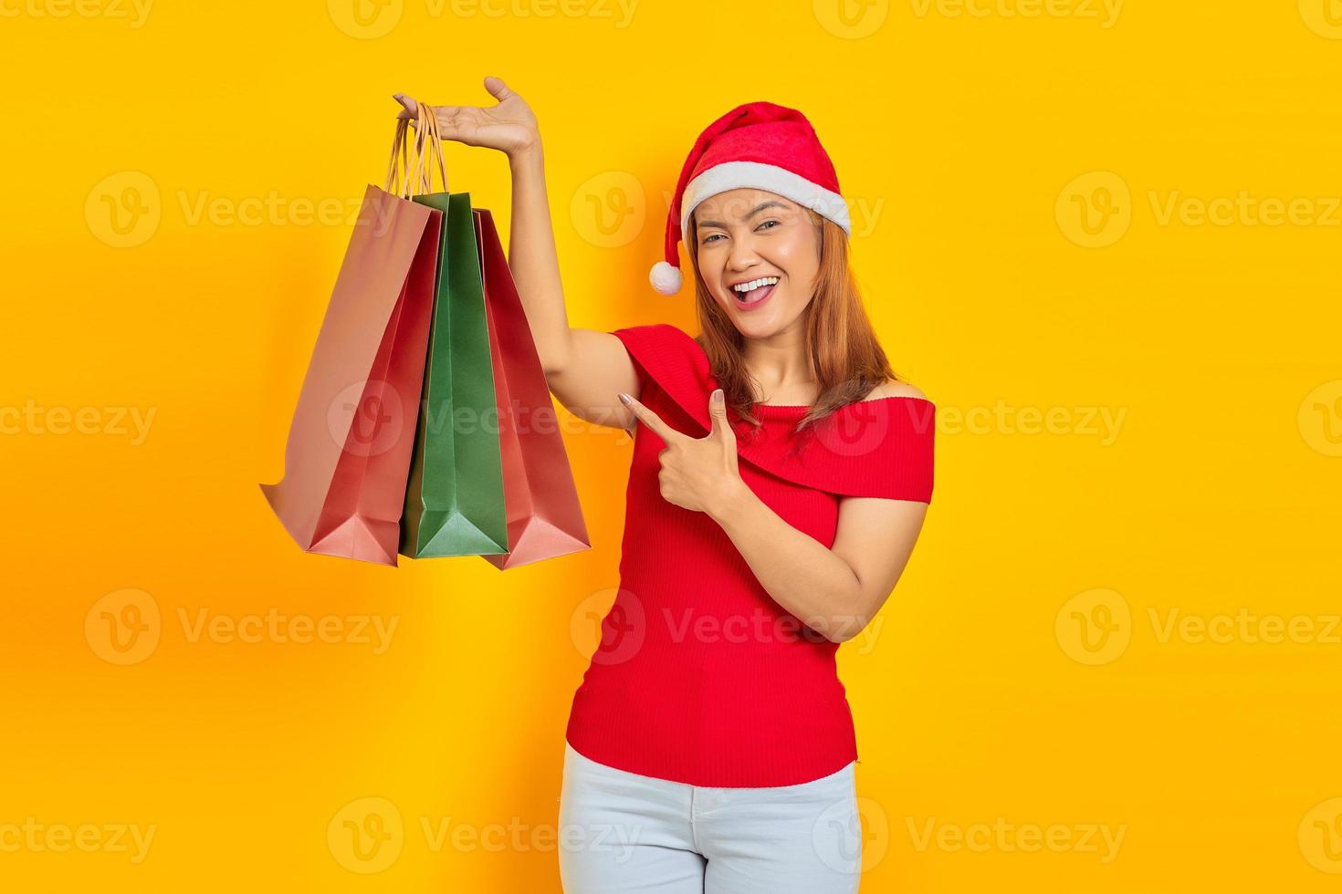 Alegre joven asiática en santa claus hat señalar con el dedo a la bolsa de compras sobre fondo amarillo foto