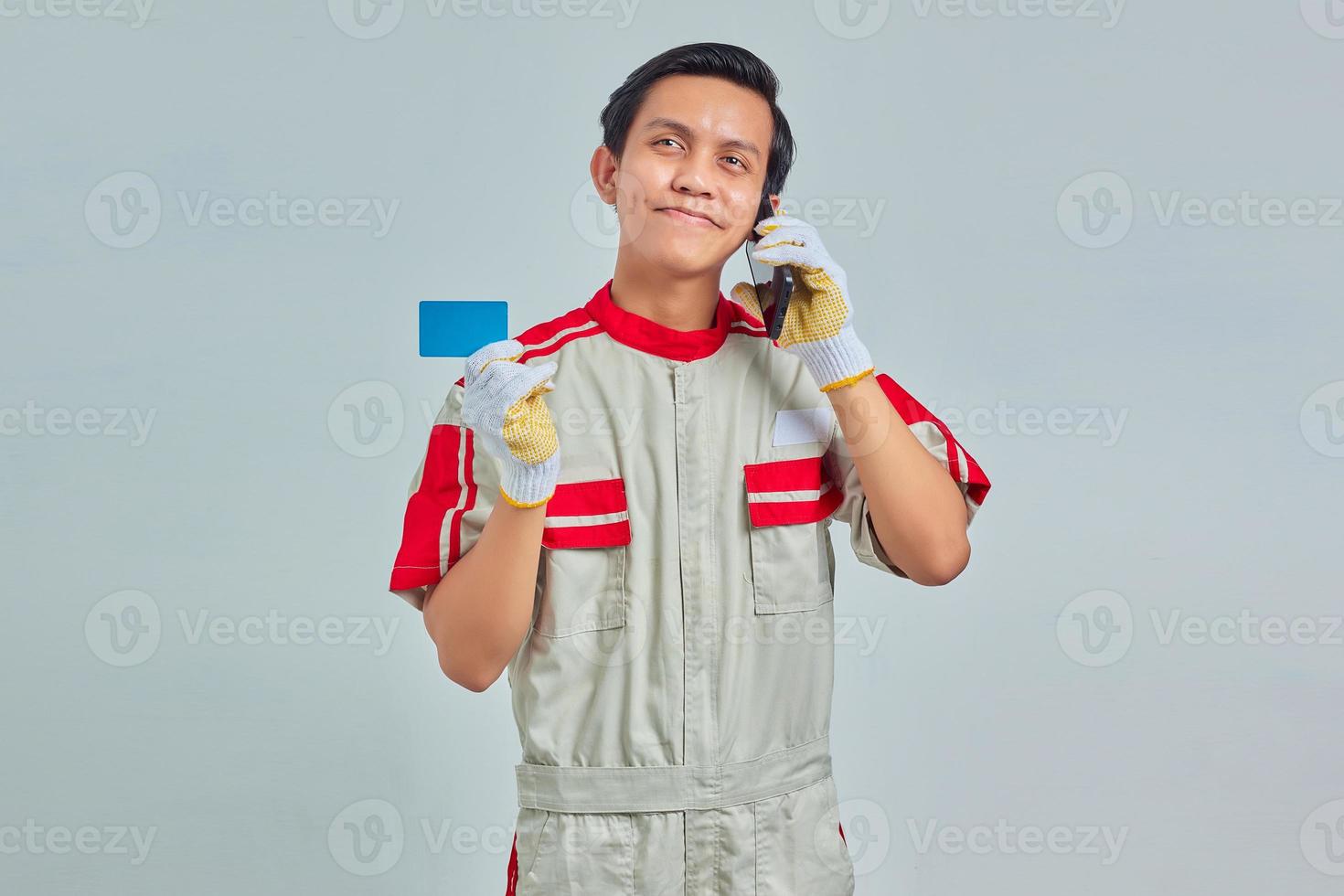 Retrato de sonriente joven mecánico mostrando tarjeta de crédito y hablando por teléfono celular sobre fondo gris foto