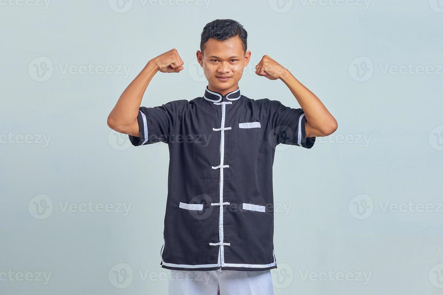 Portrait of Asian young man wearing taekwondo kimono showing muscles in hands on gray background photo