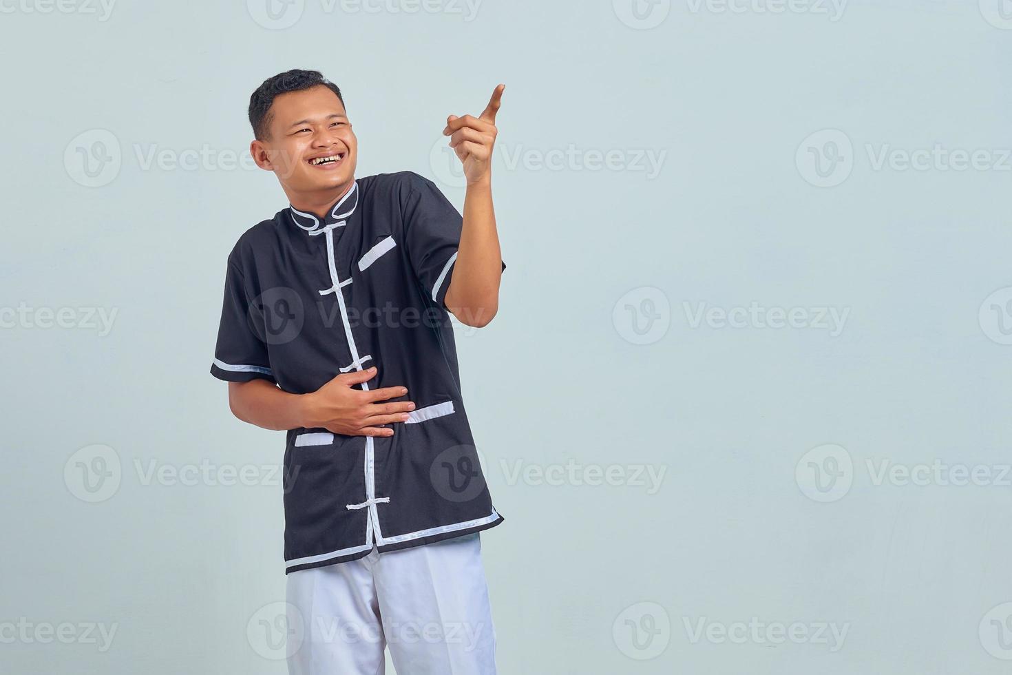 retrato, de, asiático, joven, llevando, uniforme de karate, reír, en, divertido, loco, broma, con, manos, cuerpo, encima, fondo gris foto