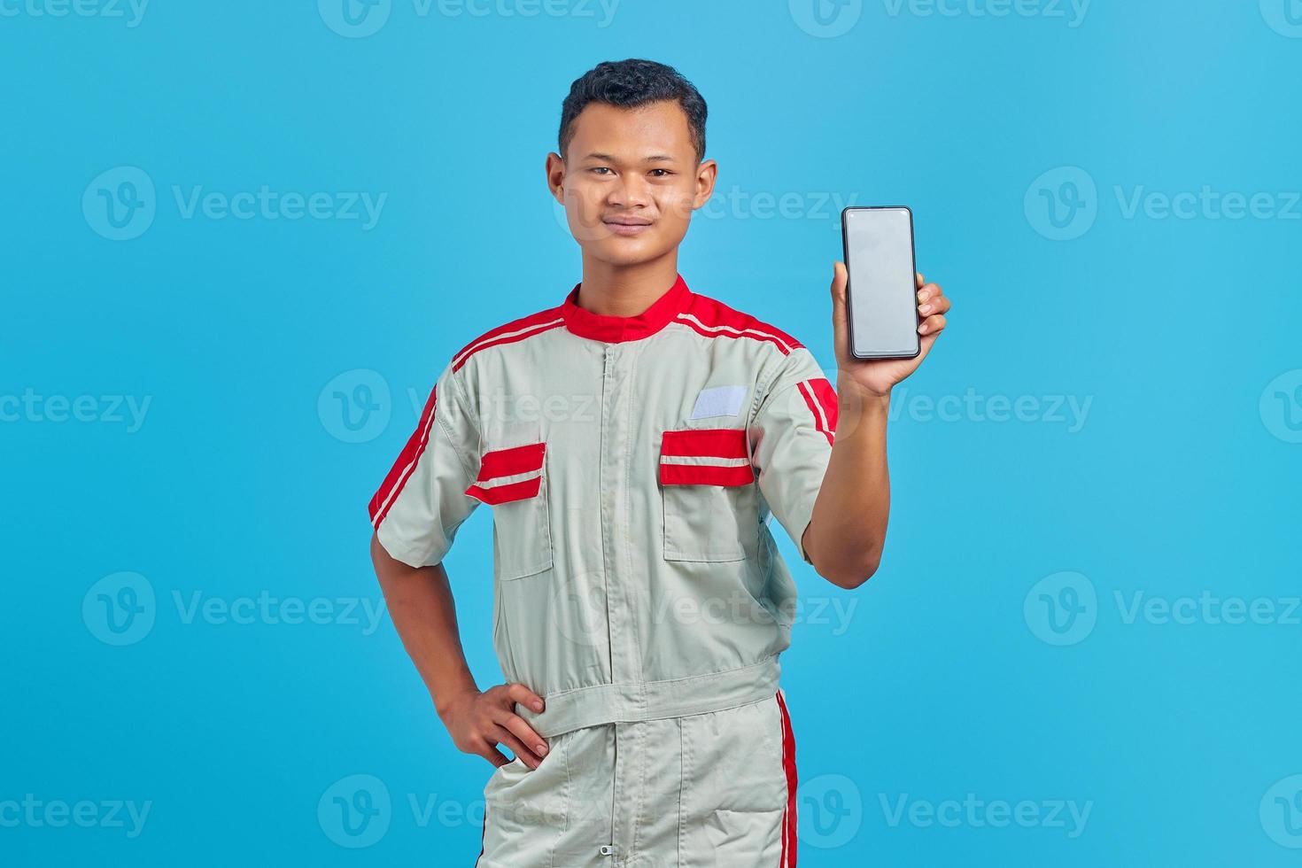 Portrait of smiling young mechanic showing blank smartphone screen in hand isolated on blue background photo