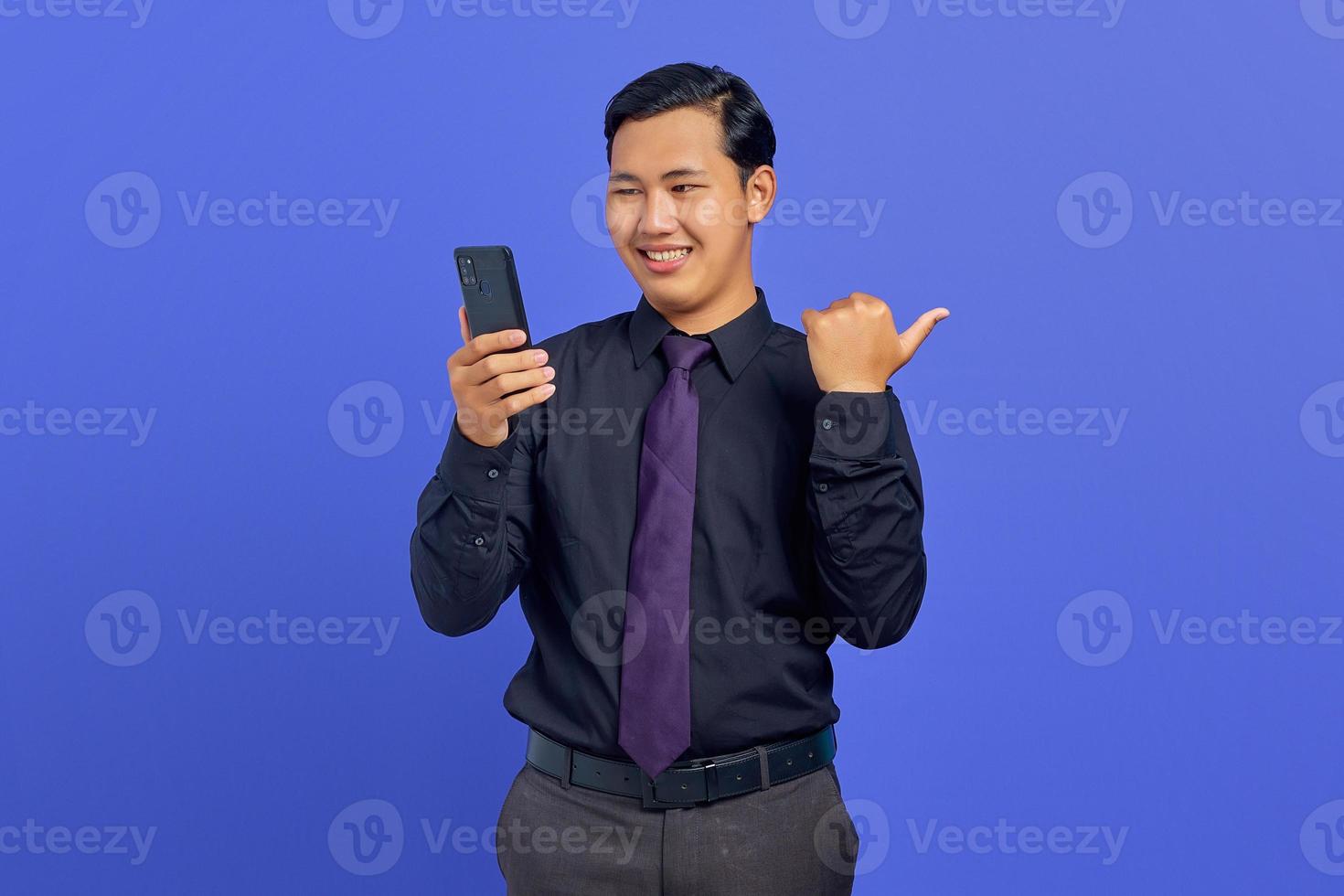 Cheerful handsome young businessman holding smartphone and pointing to copy space on purple background photo