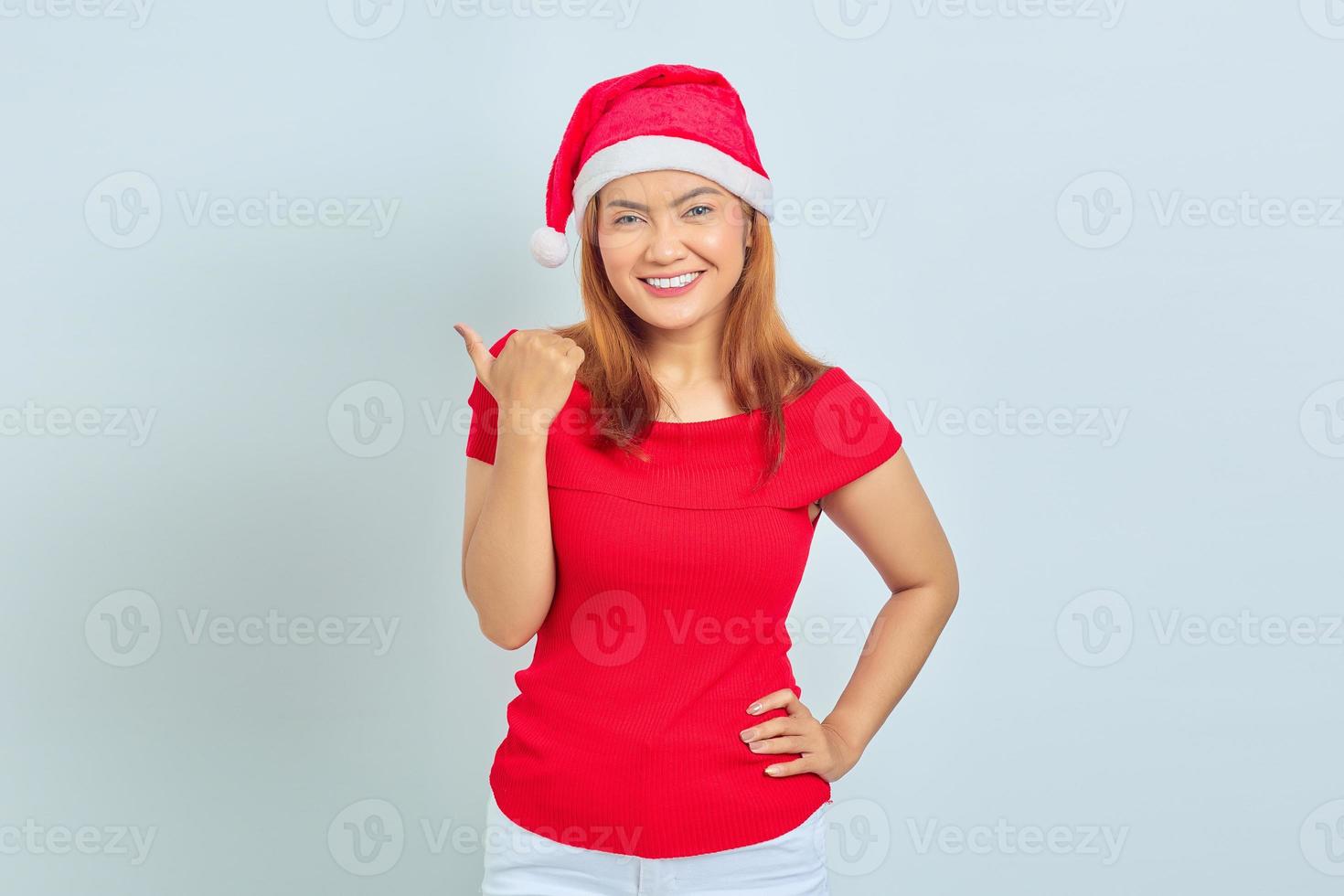 Photo of beautiful Asian girl in christmas hat with smiling facial expression and pointing aside on white background