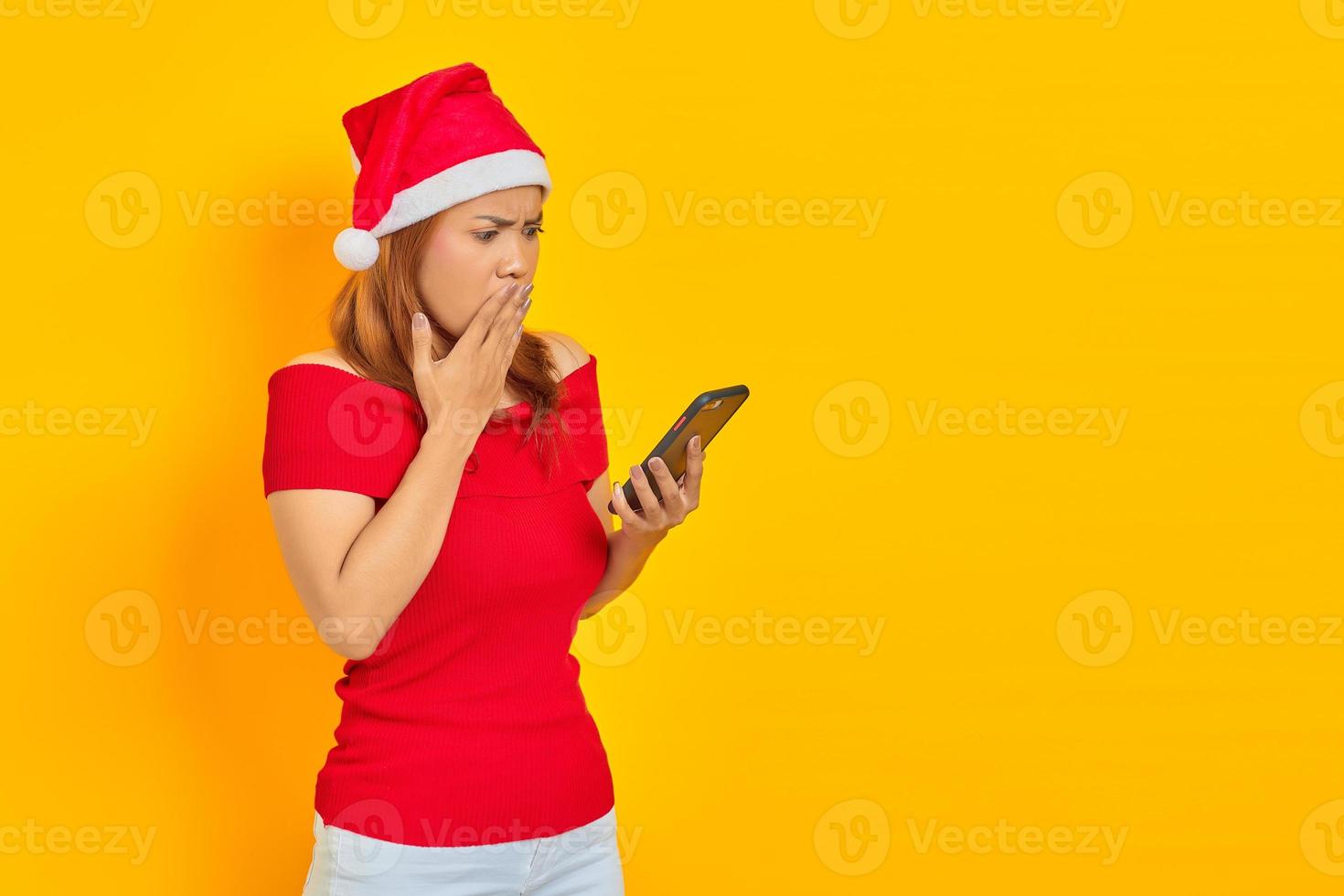 Sorprendido joven asiática en santa claus hat mirando el teléfono móvil con la boca abierta aislado sobre fondo amarillo foto