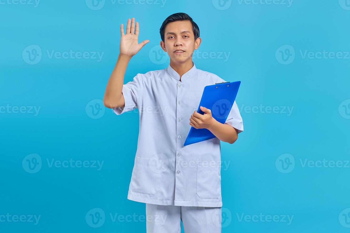 Portrait of cheerful male nurse holding clipboard standing and waving forward on blue background photo