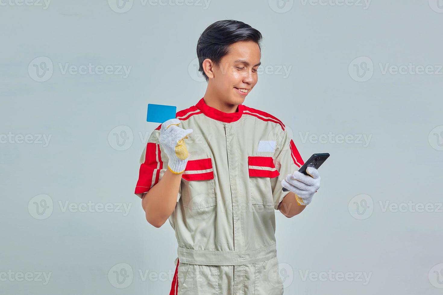 Portrait of Cheerful young mechanic showing credit card and using smartphone on gray background photo