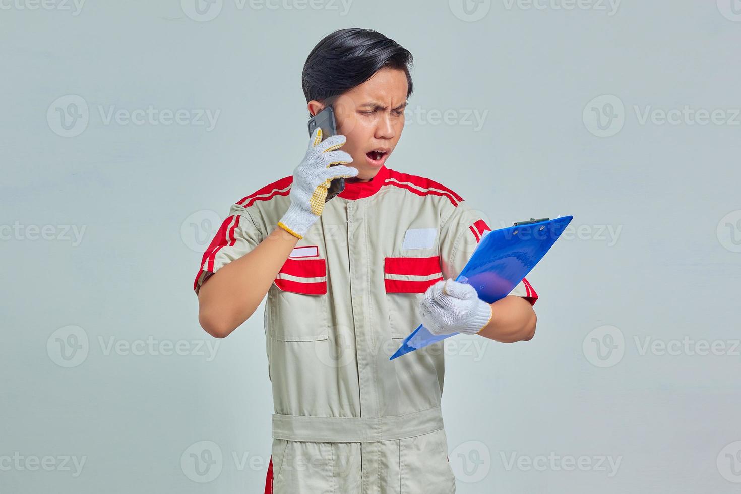 Portrait of young Asian mechanic talking on phone and holding clipboard with surprised expression isolated on gray background photo