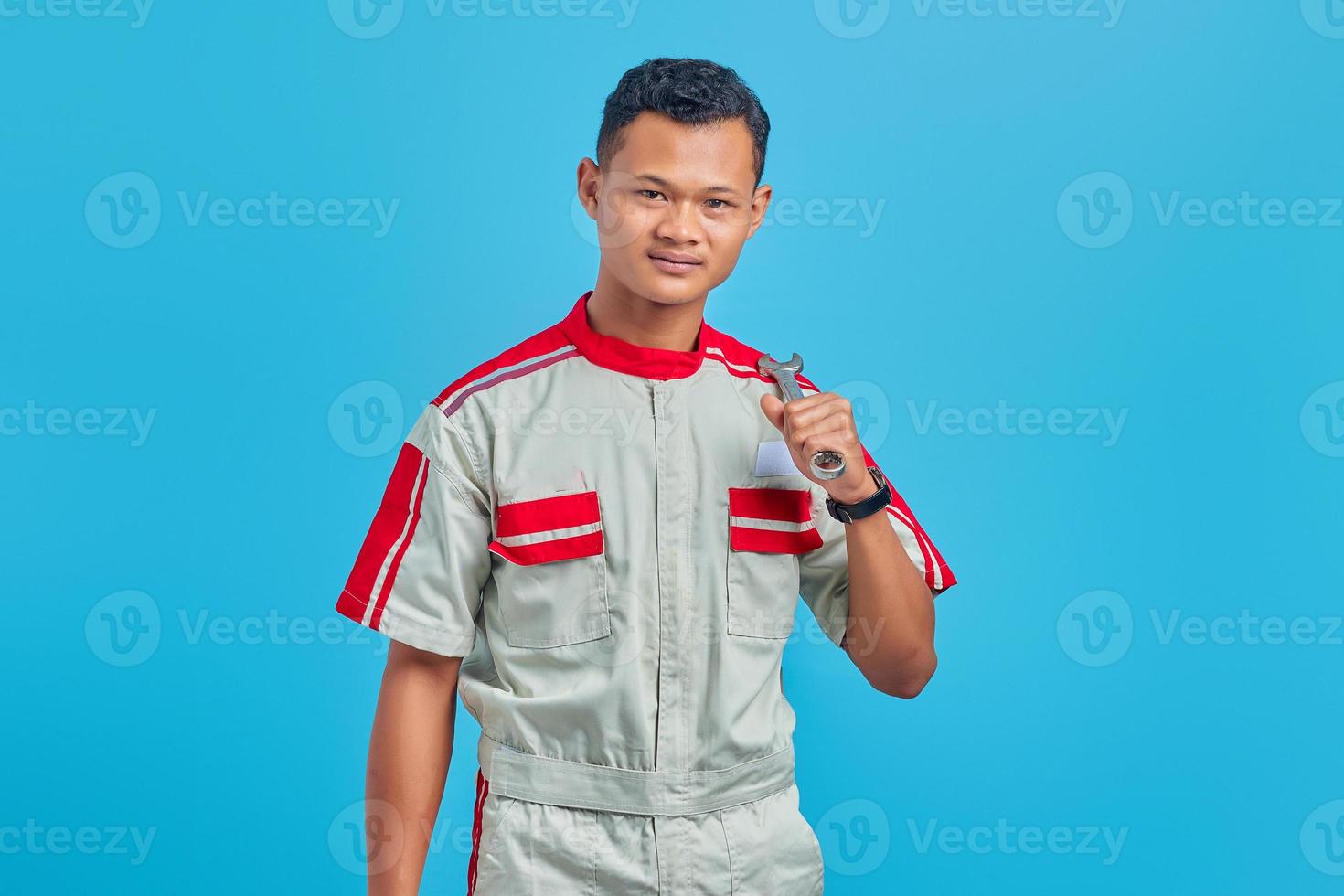 Image of smiling Asian handsome man wearing mechanical uniform holding wrench isolated on blue background photo