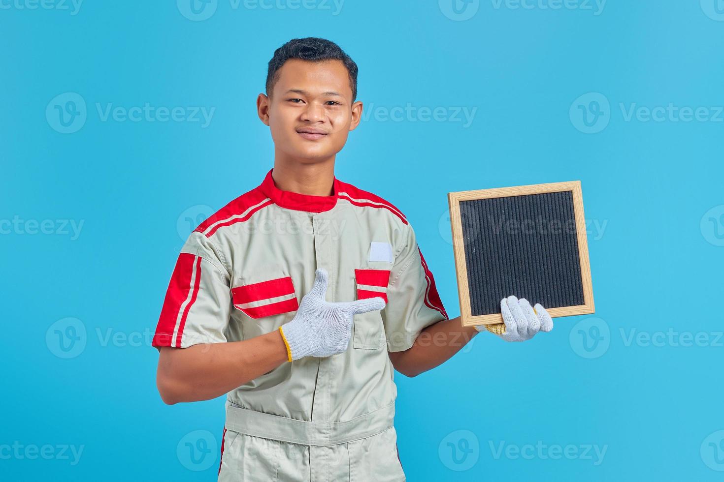 Retrato de joven alegre mecánico asiático apuntando a la placa en blanco con el dedo sobre fondo azul. foto