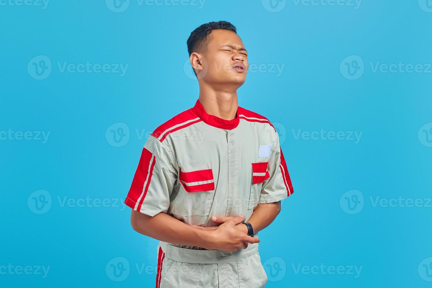 Retrato de joven mecánico asiático en dolor con las manos en el estómago sobre fondo azul. foto