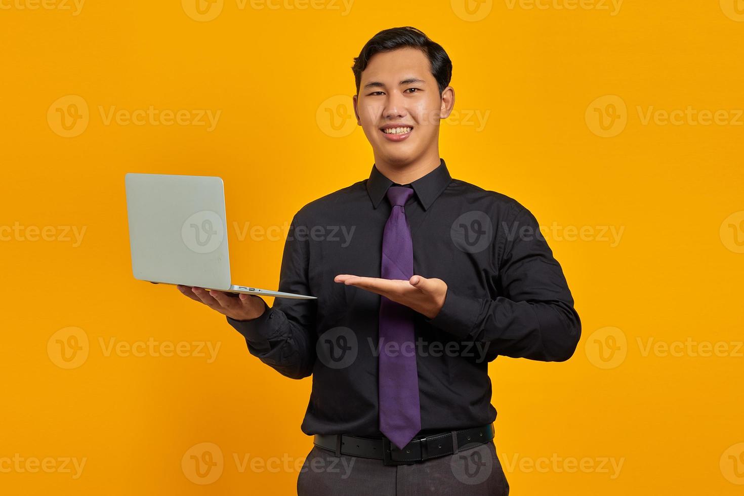 Portrait of smiling young Asian businessman showing palms and holding laptop on yellow background photo