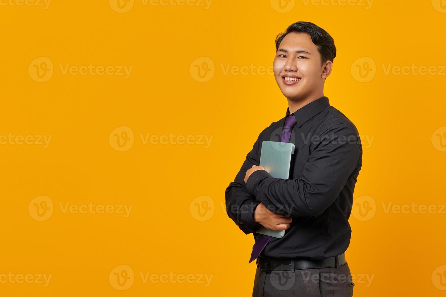 Smiling handsome young businessman hugging laptop and looking at camera on yellow background photo