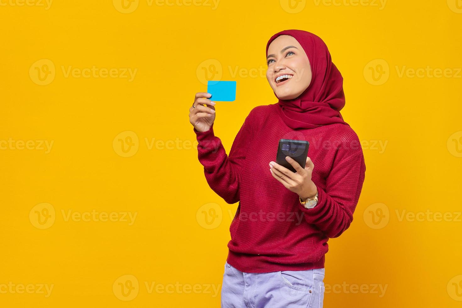 retrato, de, pensativo, joven, mujer asiática, mirar hacia arriba, tenencia, tarjeta de crédito, y, teléfono móvil, en, fondo amarillo foto