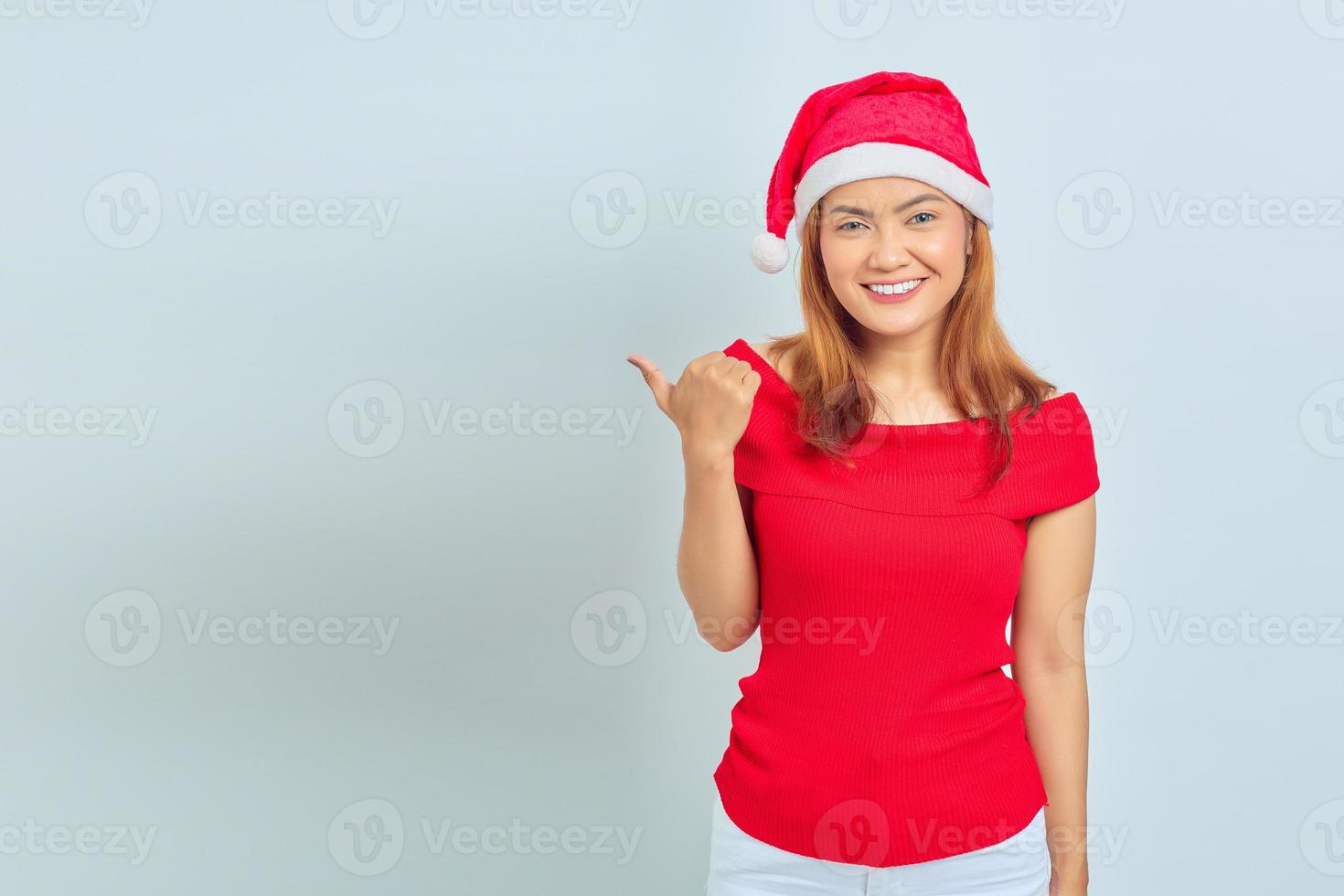 Photo of beautiful Asian girl in christmas hat with smiling facial expression and pointing aside on white background