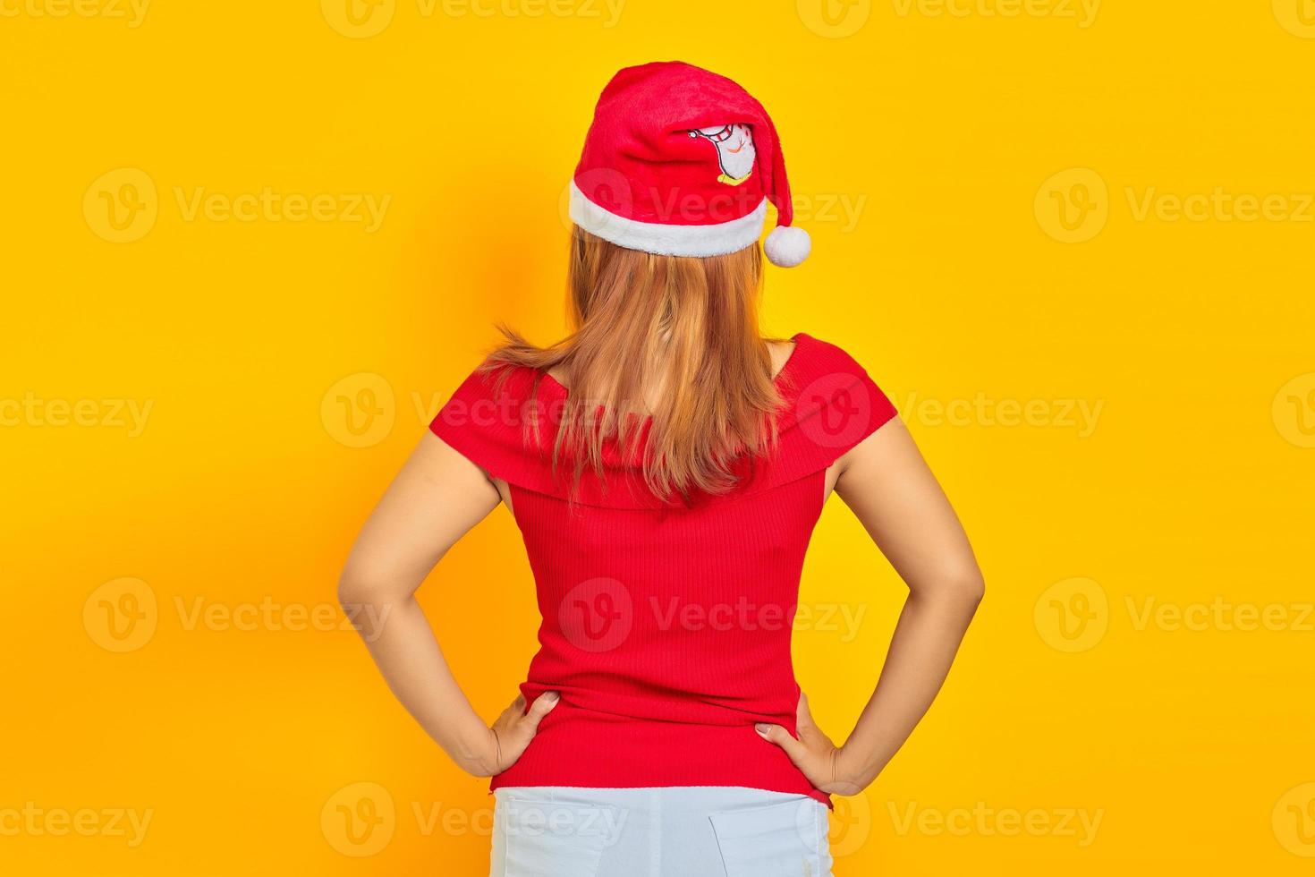 Young woman wearing Christmas hat in back position holding hands on waist confidently on yellow background photo