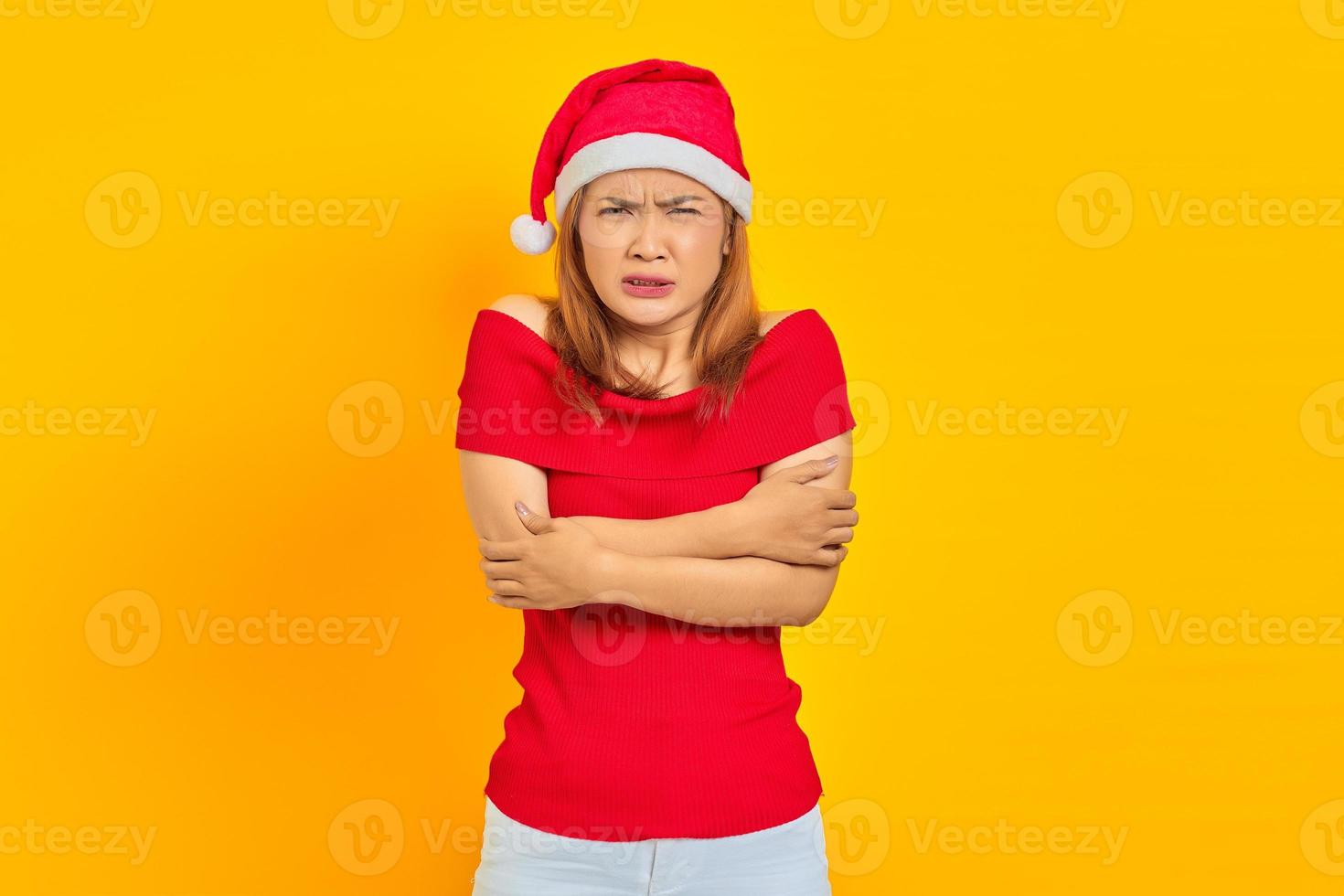 Portrait of young Asian woman wearing Christmas hat and showing freeze gesture on yellow background photo