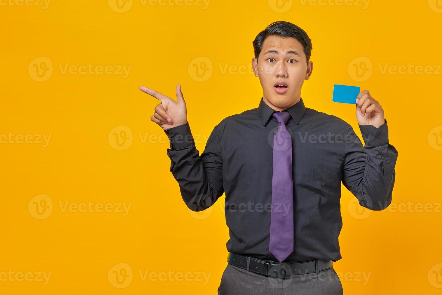Shocked asian businessman holding a credit card while pointing finger at copy space on yellow background photo
