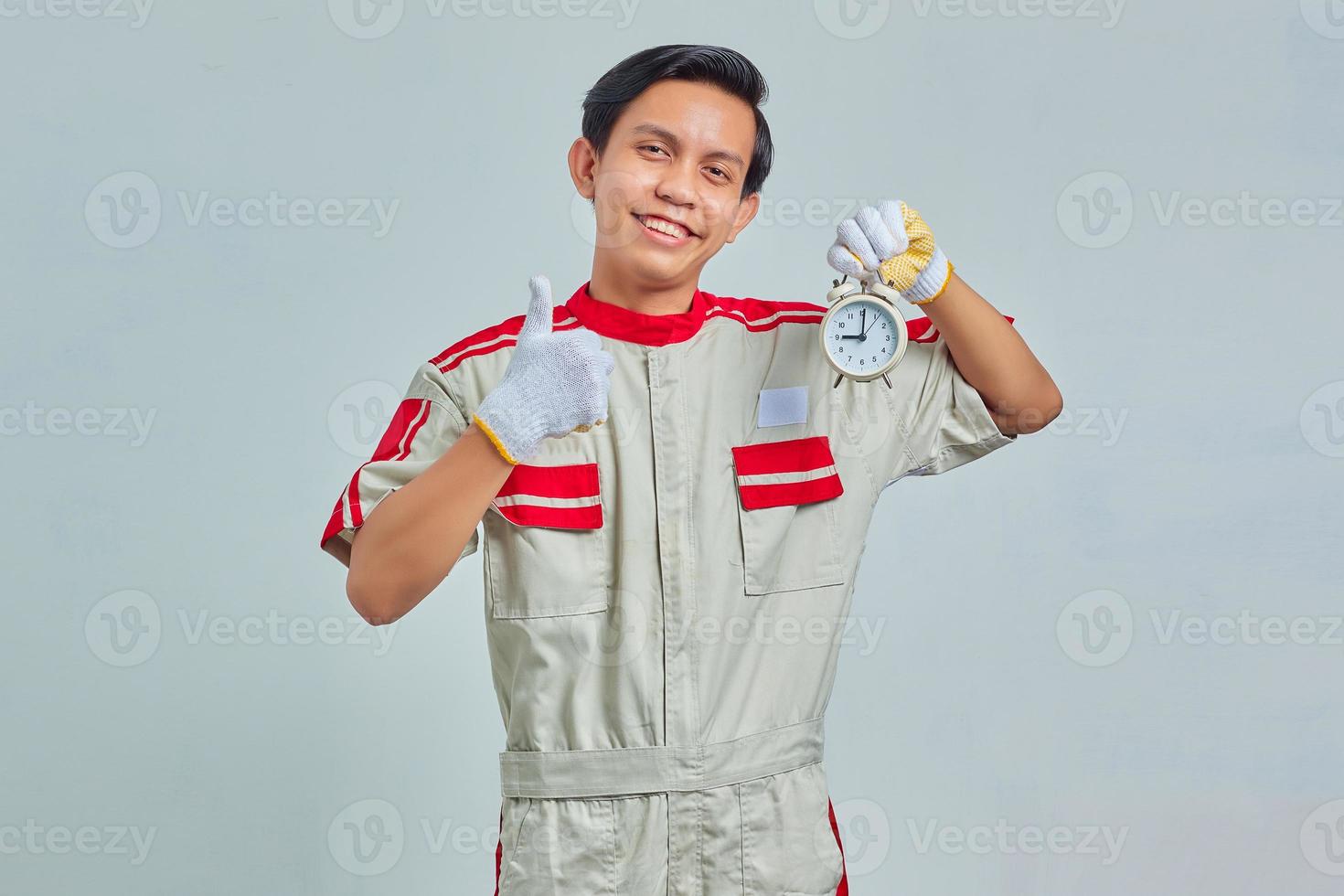 Portrait of happy handsome man wearing mechanical uniform holding alarm clock and thumb up on gray background photo