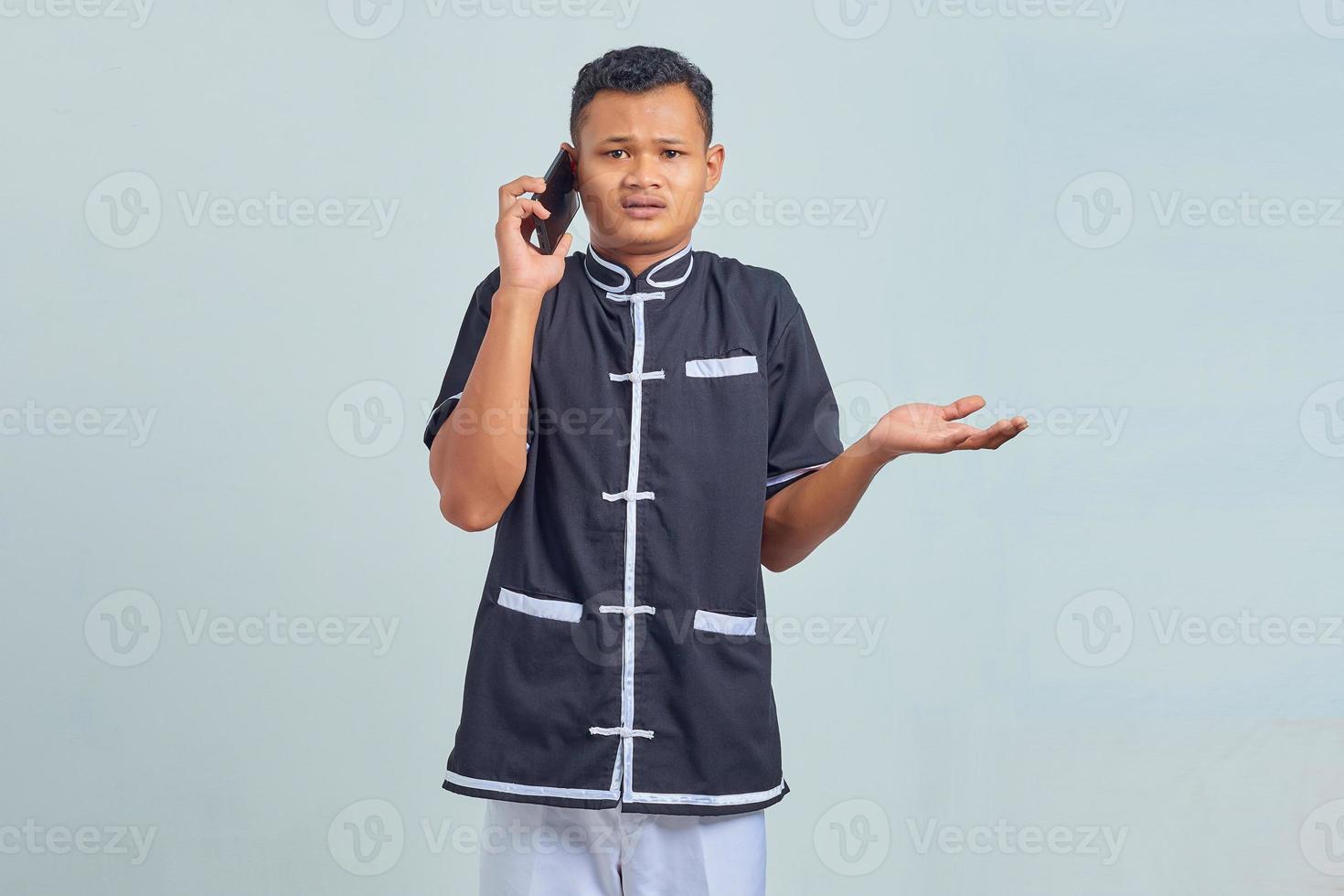 Portrait of confused asian young man wearing karate uniform talking on smartphone on gray background photo