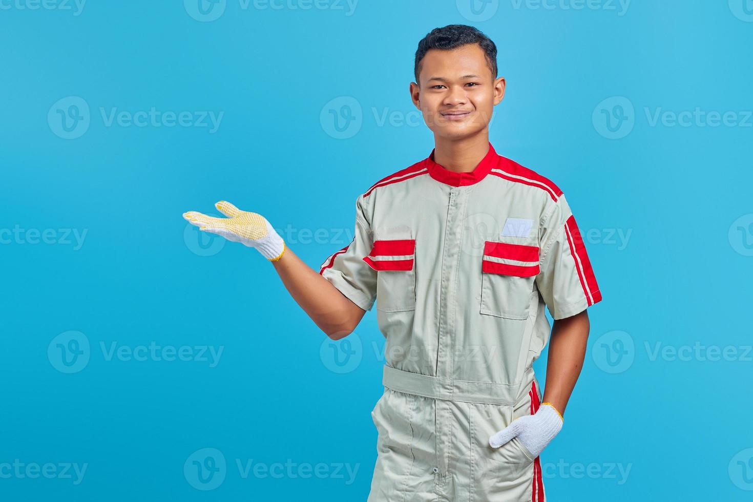 Retrato de smilling joven mecánico asiático mostrando copia espacio en palmas sobre fondo azul. foto