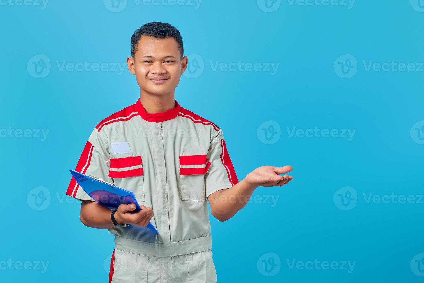 Portrait of excited young mechanic holding clipboard and pointing with palms pointing sideways isolated on blue background photo