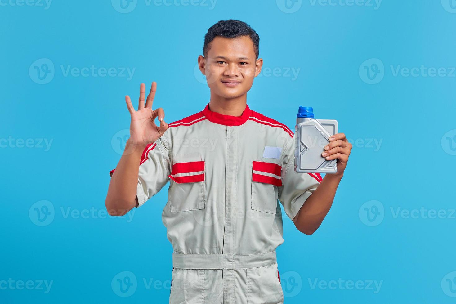 Retrato de sonriente joven mecánico asiático sosteniendo una botella de plástico de aceite de motor y mostrando aprobación con el pulgar hacia arriba sobre fondo azul. foto