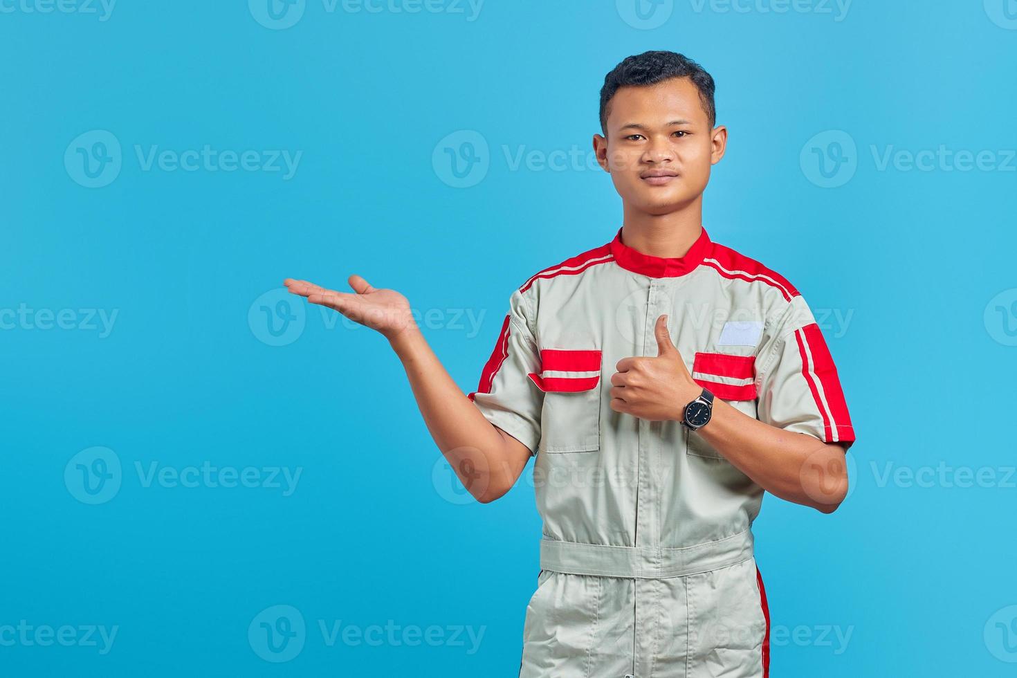 Portrait of smilling young Asian mechanic  showing palm hands and thumbs up over blue background photo