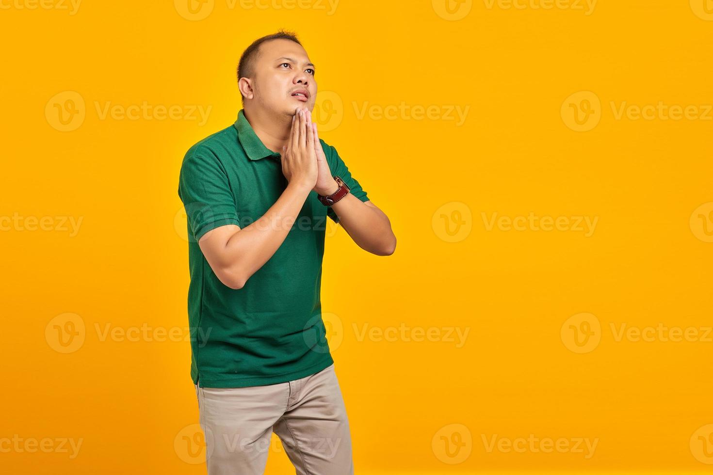 Retrato de un joven asiático suplicando y orando con las manos junto con la expresión de esperanza en la cara muy emocional y preocupada sobre fondo amarillo foto