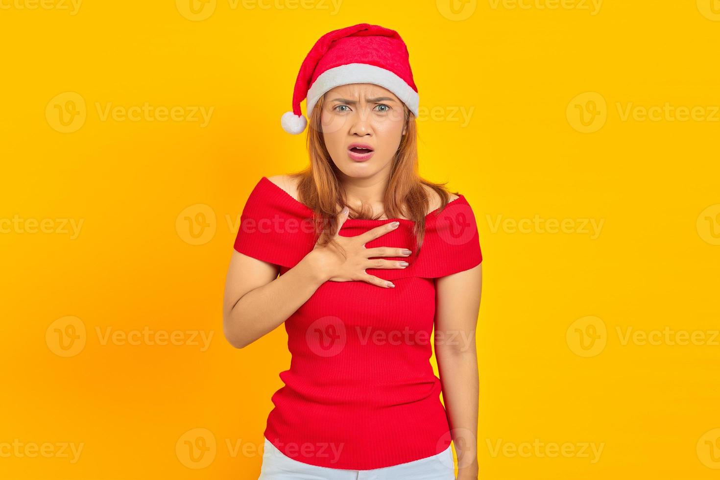 Shocked young Asian woman wearing Christmas hat and put hands chest on yellow background photo