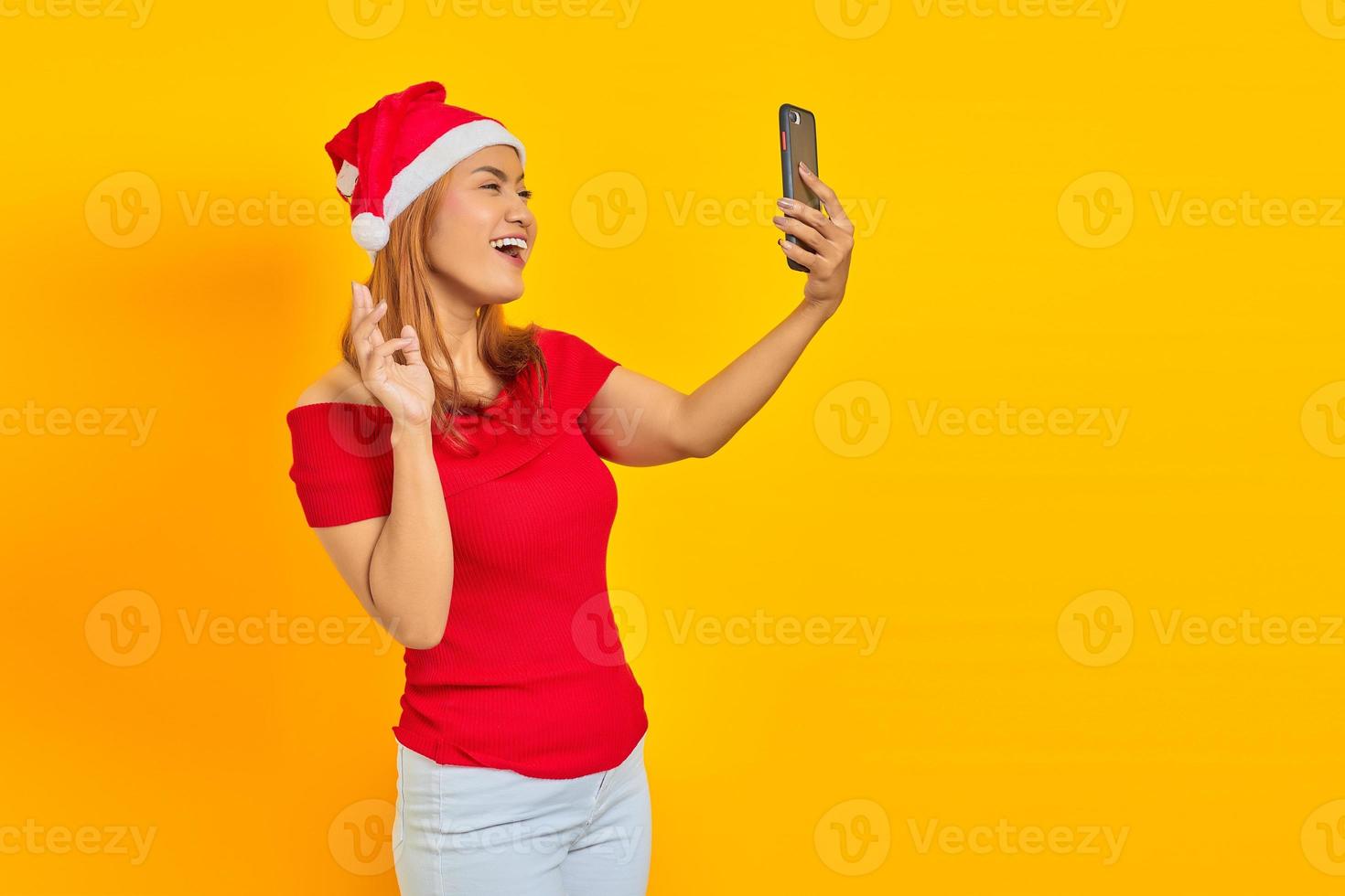 Cheerful young Asian woman wearing Christmas hat talking a selfie with cellphone on yellow background photo