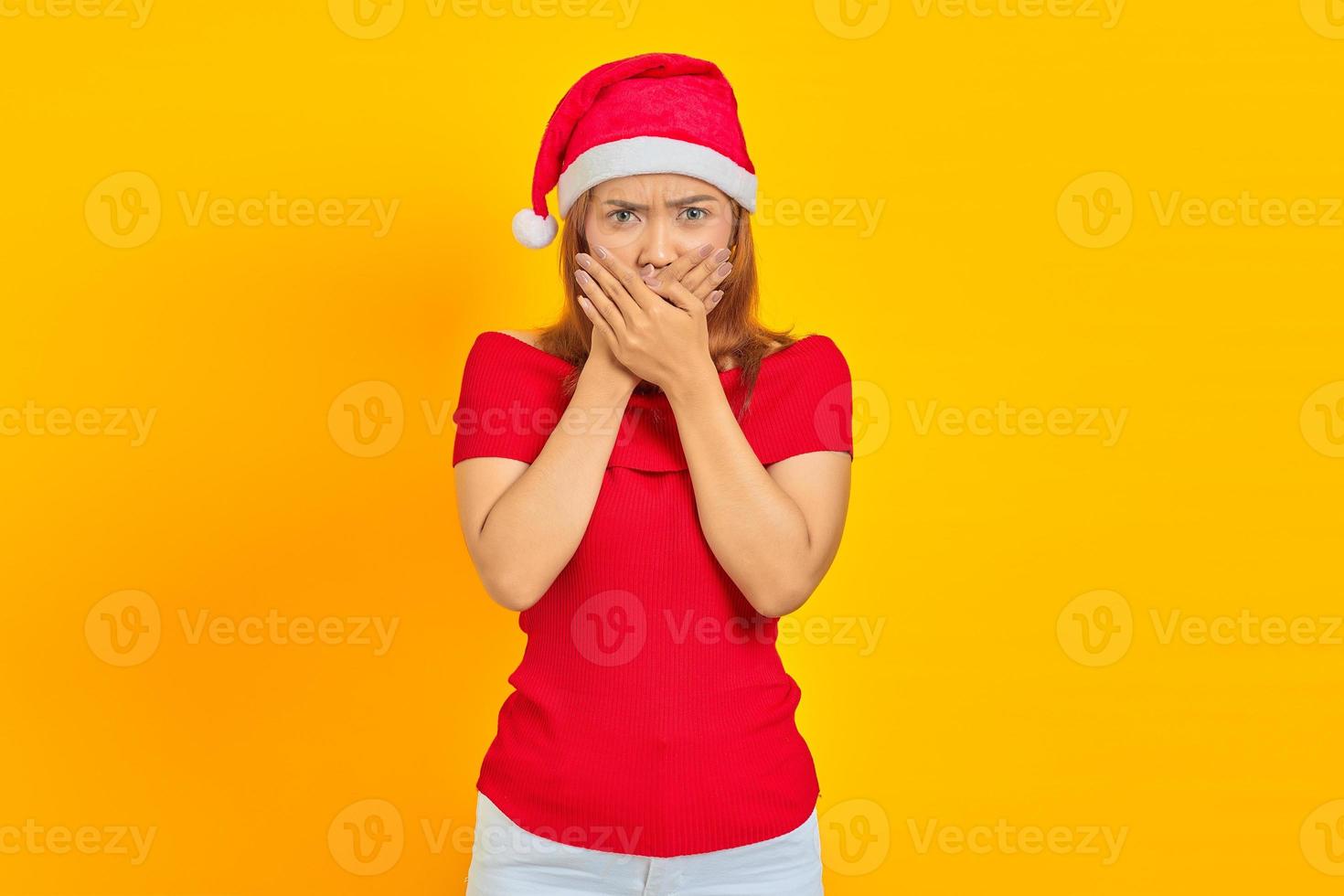 Surprised young Asian woman wearing Christmas hat covering mouth with hands on yellow background photo