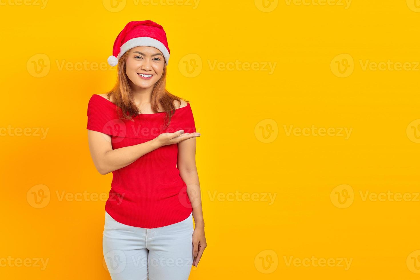 Smiling young Asian woman wearing a christmas hat and extending hand to the side inviting to come photo