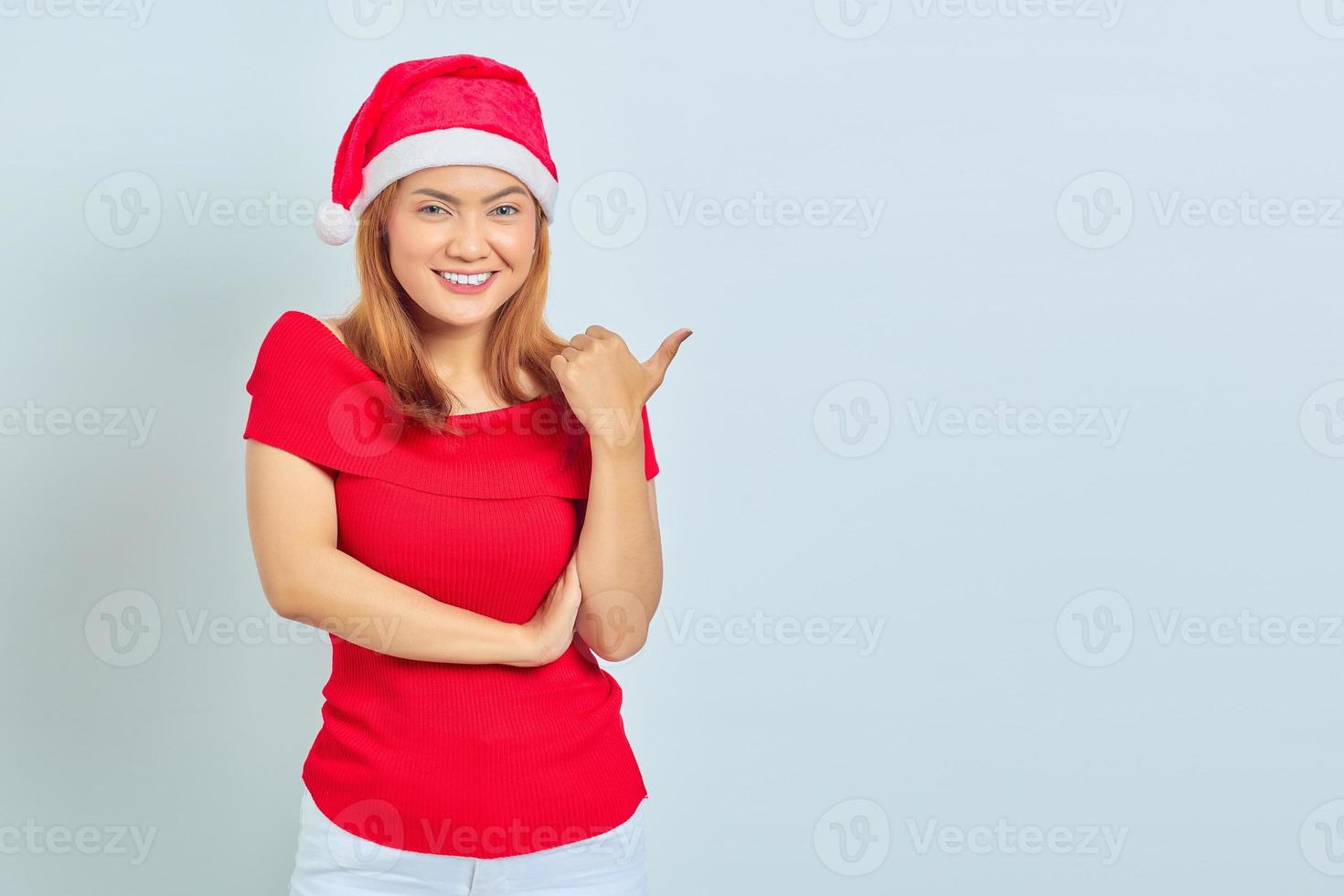 Foto de hermosa chica asiática con sombrero de navidad con expresión facial sonriente y apuntando a un lado sobre fondo blanco.