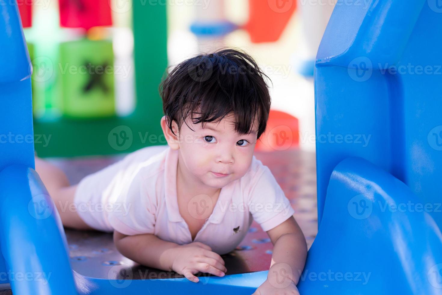 El pequeño bebé asiático juega en el equipo del patio de recreo en el calor del verano. El niño mira a la cámara y sonríe levemente. niño pequeño sudando en la cara. niño de 2 años. foto