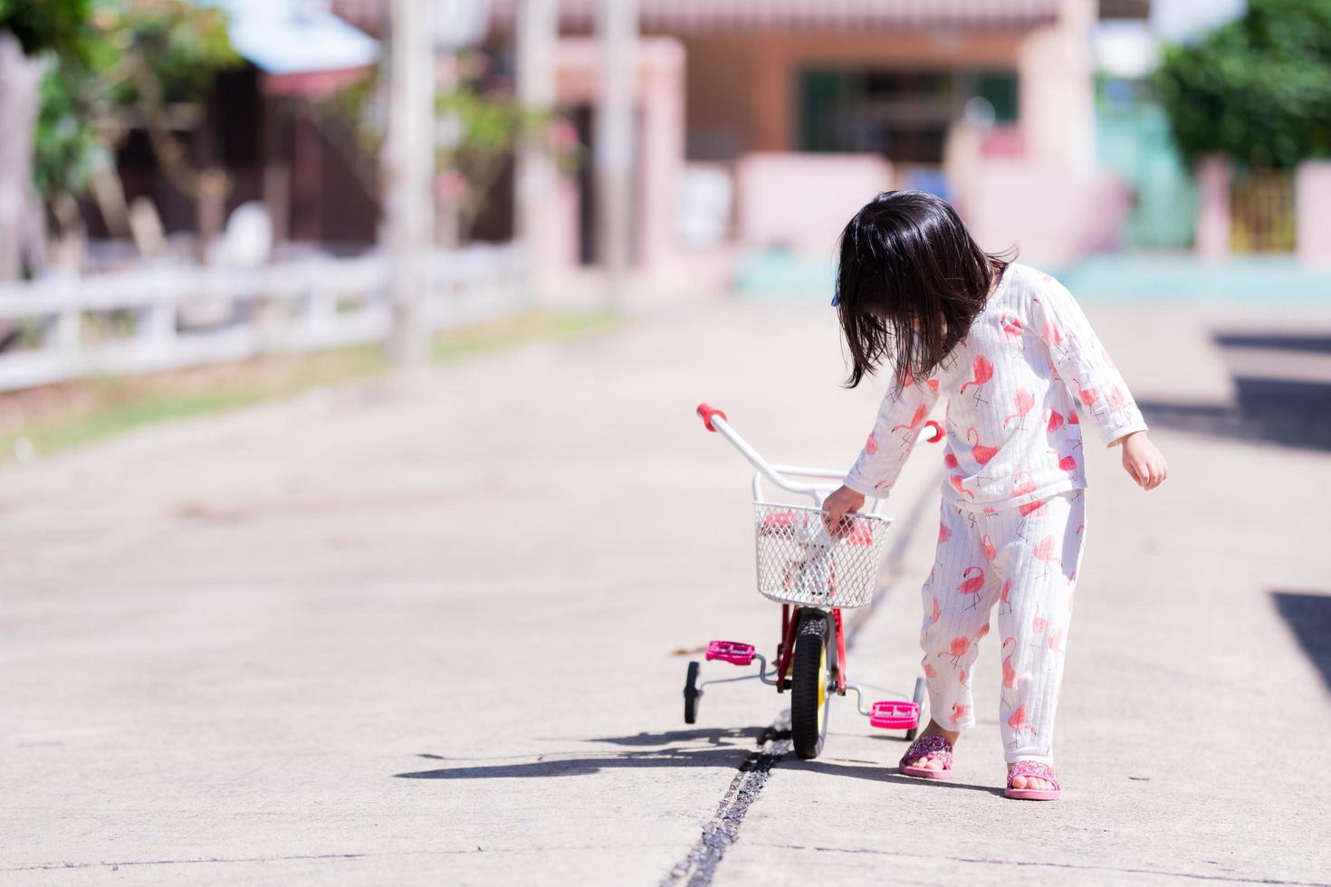 Asian girl take walk in morning on street. Child is leading red bicycle. Baby wear pajamas. Kid aged 3-4 years old. photo