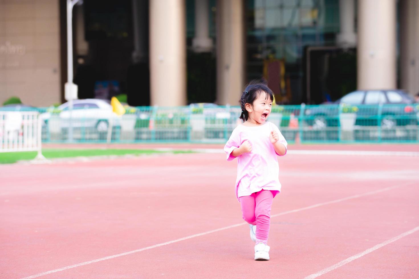 chica activa corriendo diviértete en la pista roja en el estadio. niño feliz sonriendo y riendo. ejercicio infantil. El bebé con vestido rosa tiene 3-4 años. foto