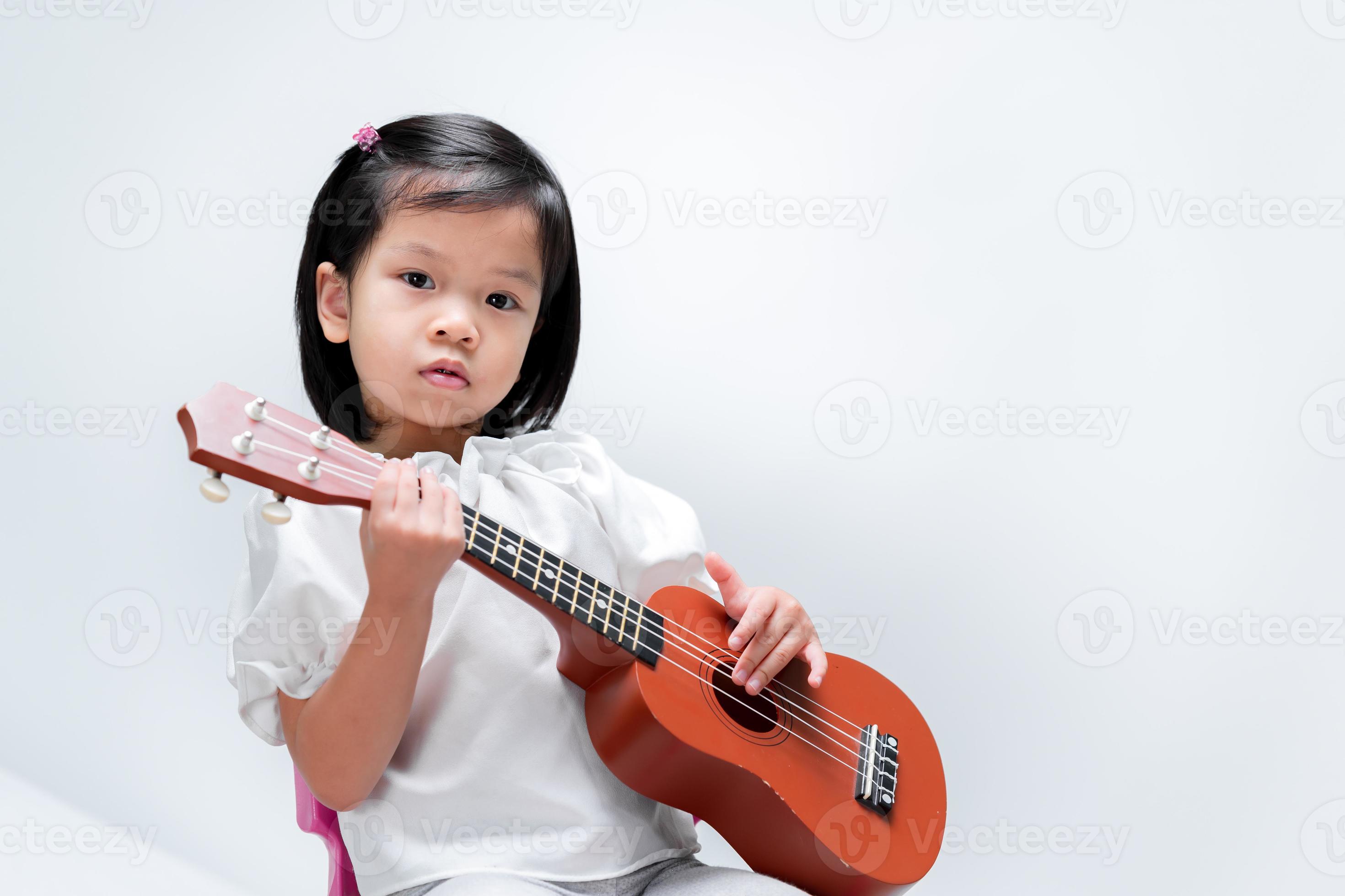 niño lindo aprende a tocar la guitarra pequeña en la escuela de música. chica se encuentra con ukeleles sobre fondo gris. foto