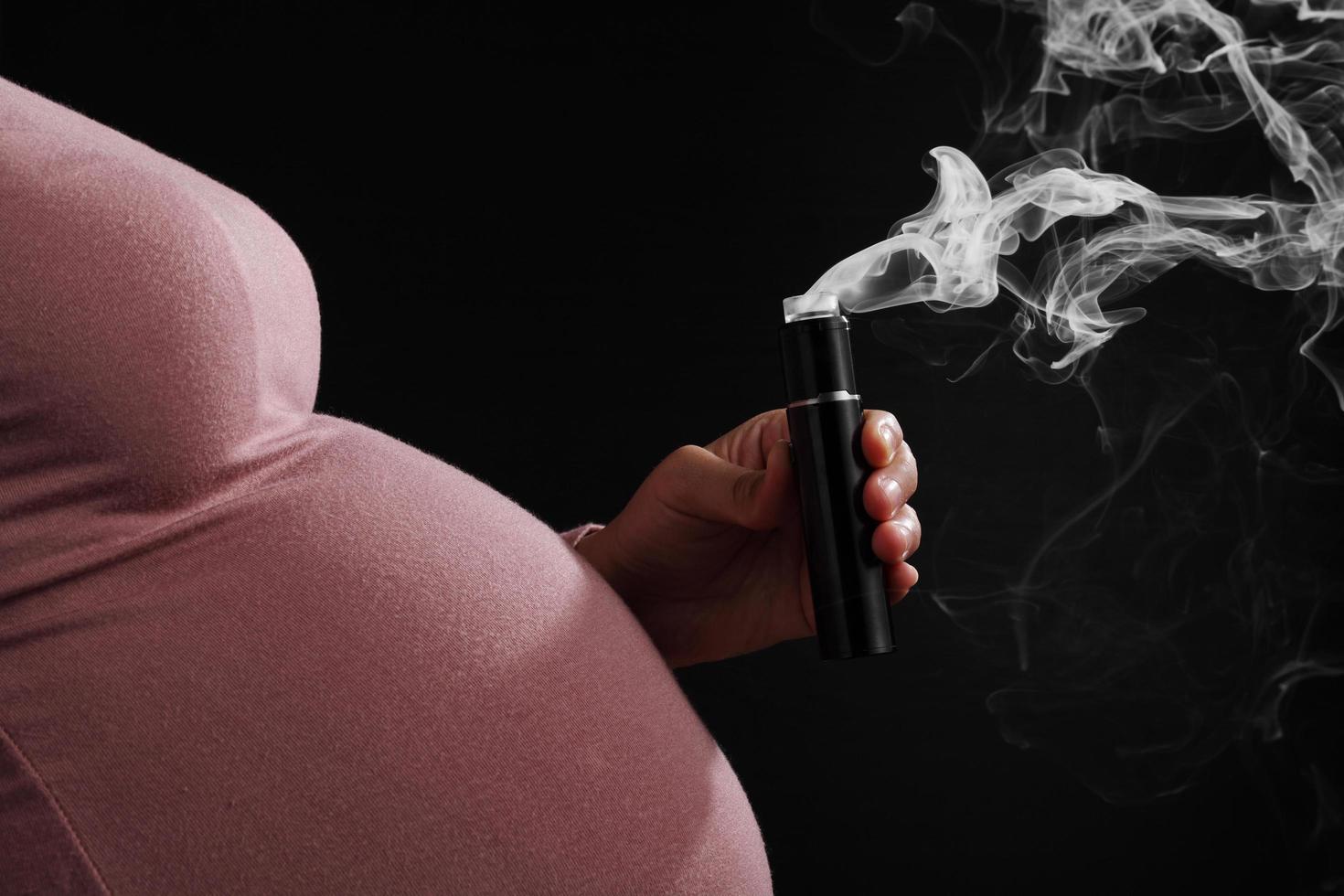 Close up pregnant woman smoking e-cigarette on black background photo