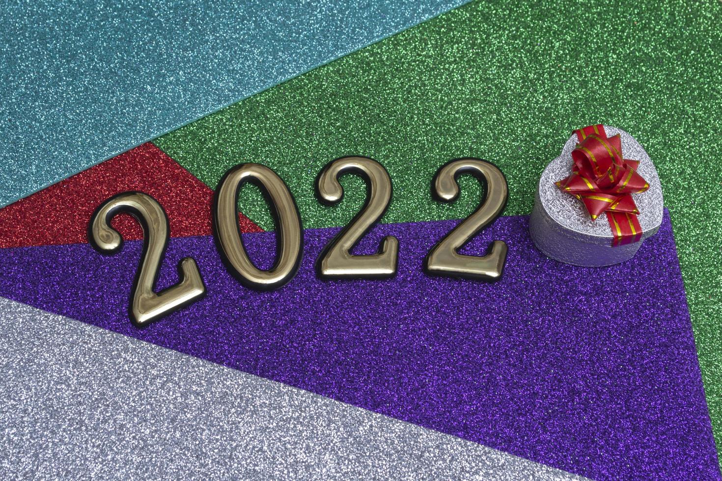 sobre un fondo de año nuevo brillante colorido, una caja de regalo plateada con un lazo rojo y números dorados 2022. concepto para la tarjeta de año nuevo foto