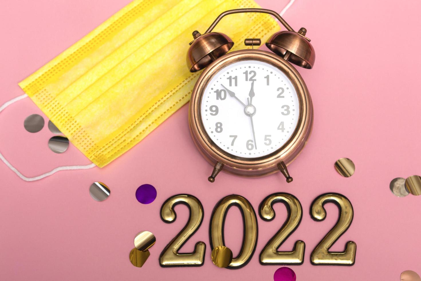 New year concept on pink background alarm clock in yellow medical mask with golden numbers 2022, close up photo