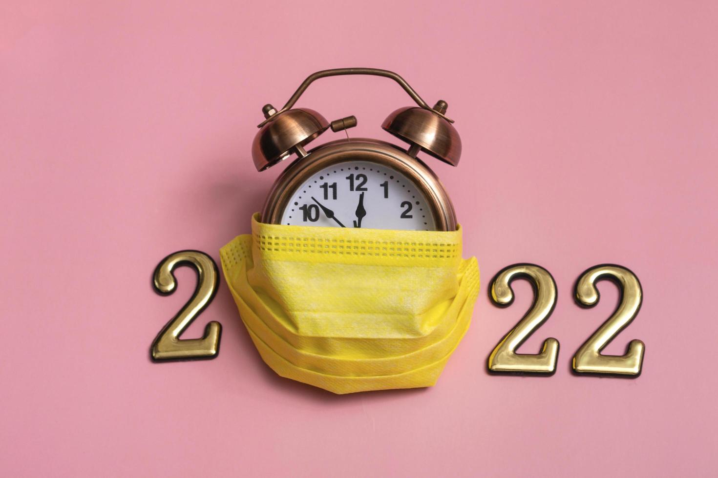 New year concept on pink background alarm clock in yellow medical mask with golden numbers 2022, photo